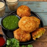 side shot of potato bread roll stacked on a black ceramic plate with green chutney