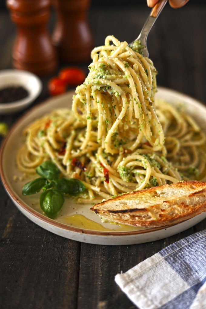 Side view of broccoli pesto pasta being lifted off plate by fork on wooden table with bread on plate.